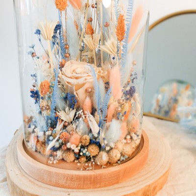 Cloche en verre et base en bois contenant des fleurs séchées blanches, pêche et bleu ciel. Grand format. Collection Pêche Bleue