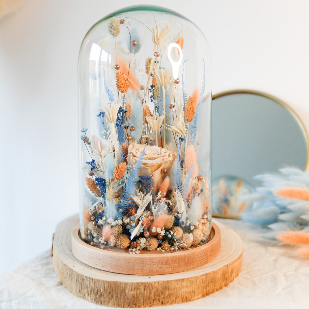 Cloche en verre et base en bois contenant des fleurs séchées blanches, pêche et bleu ciel. Grand format. Collection Pêche Bleue